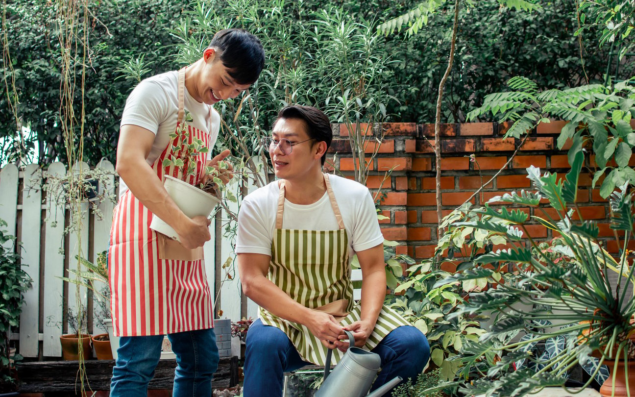 Asian handsome male LBGT couple wearing apron, sitting in outdoor garden together, doing activity as hobby in free time, helping, taking care and watering plants with refreshment, happiness and love