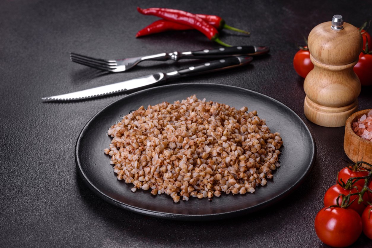 Delicious fresh boiled buckwheat porridge with vegetables and spices on a black plate against a dark concrete background