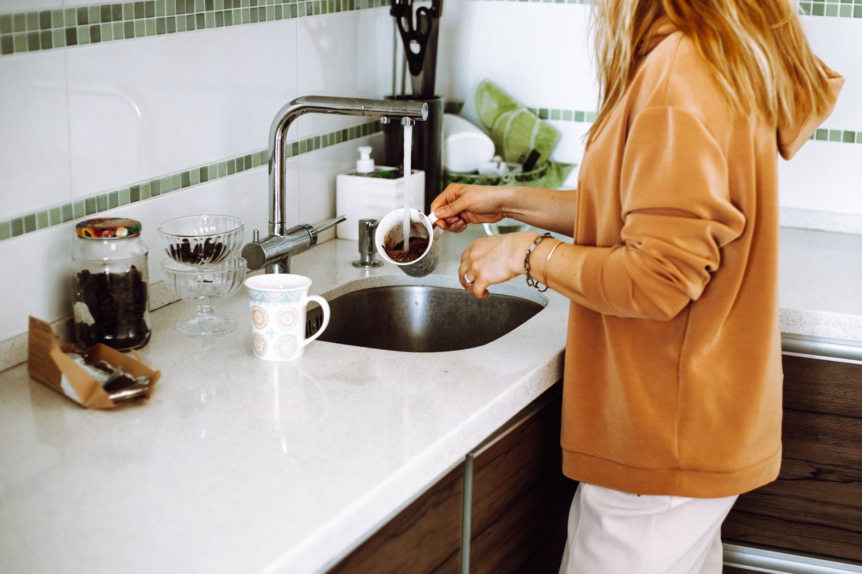 Cropped photo of young woman with wavy fair hair wearing brown sweatshirt, washing cup from coffee under open water.