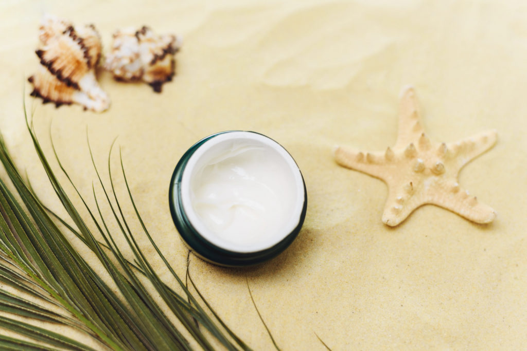 White cosmetic jar with sunscreen on textured sand background with seashells and palm leaf.