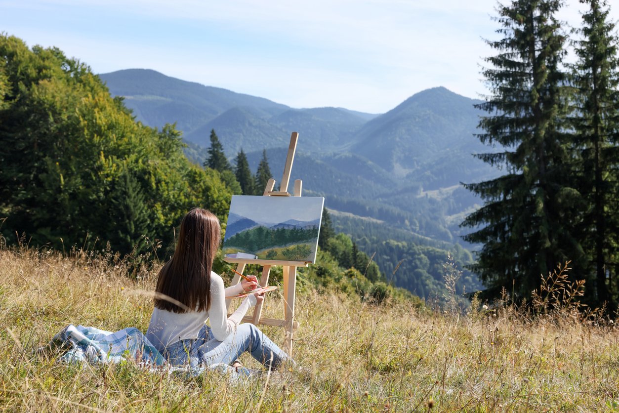 Young woman drawing on easel in mountains, back view. Space for text