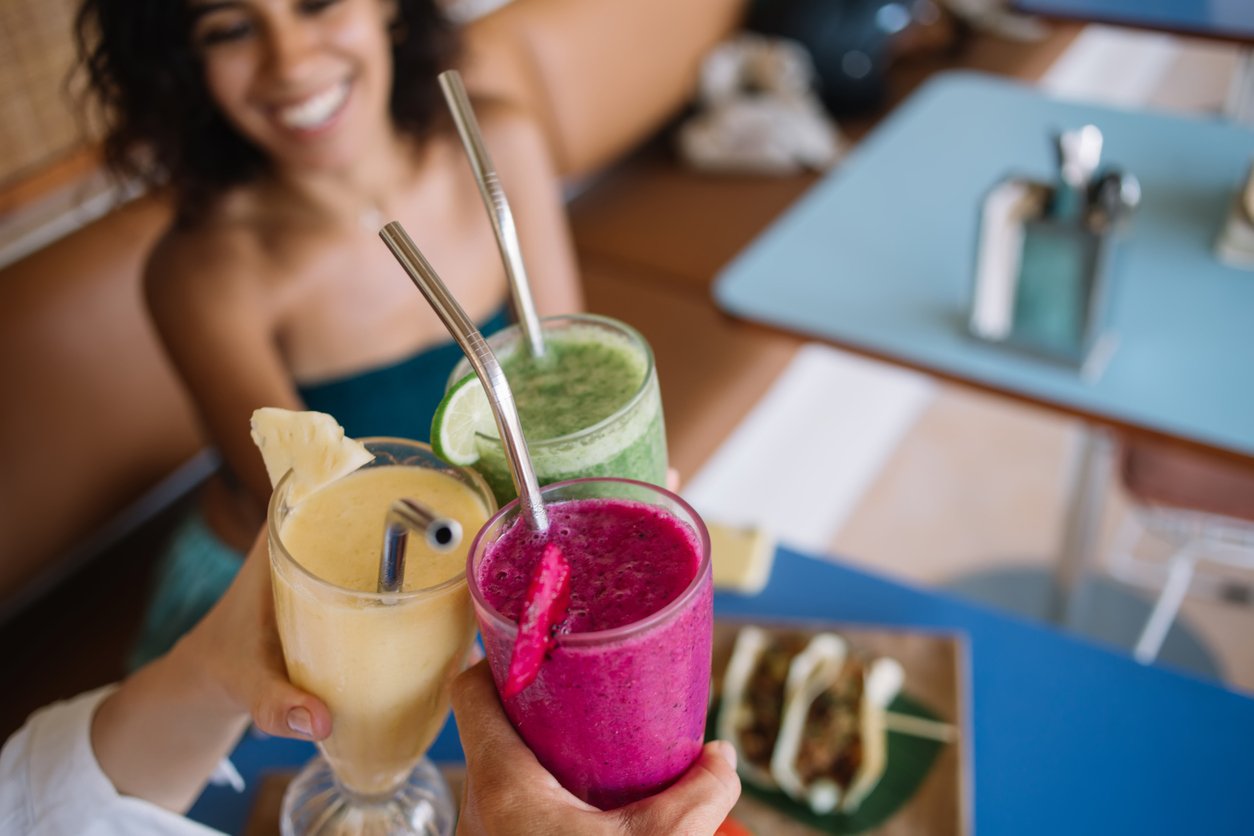 Cropped image of cafeteria customers holding smoothie cocktails and gesturing cheers during friendly meeting in bistro, selective focus on colorful milkshakes with mixed ripe fruits and fresh veggies