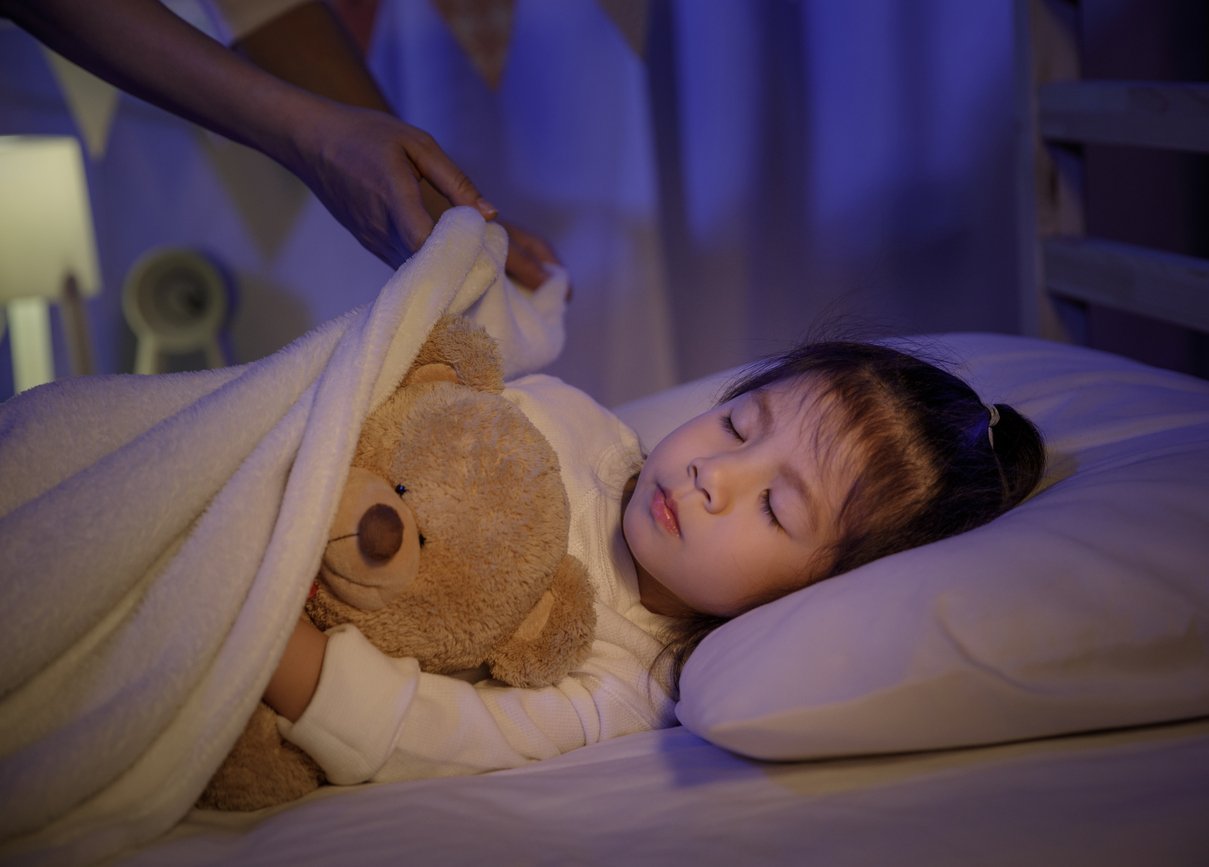 Mother putting blanket on and stroking head for her little girl daughter on bed in a dark bedroom at night, Child asian girl hug teddy bear, Comfortable children at home concept