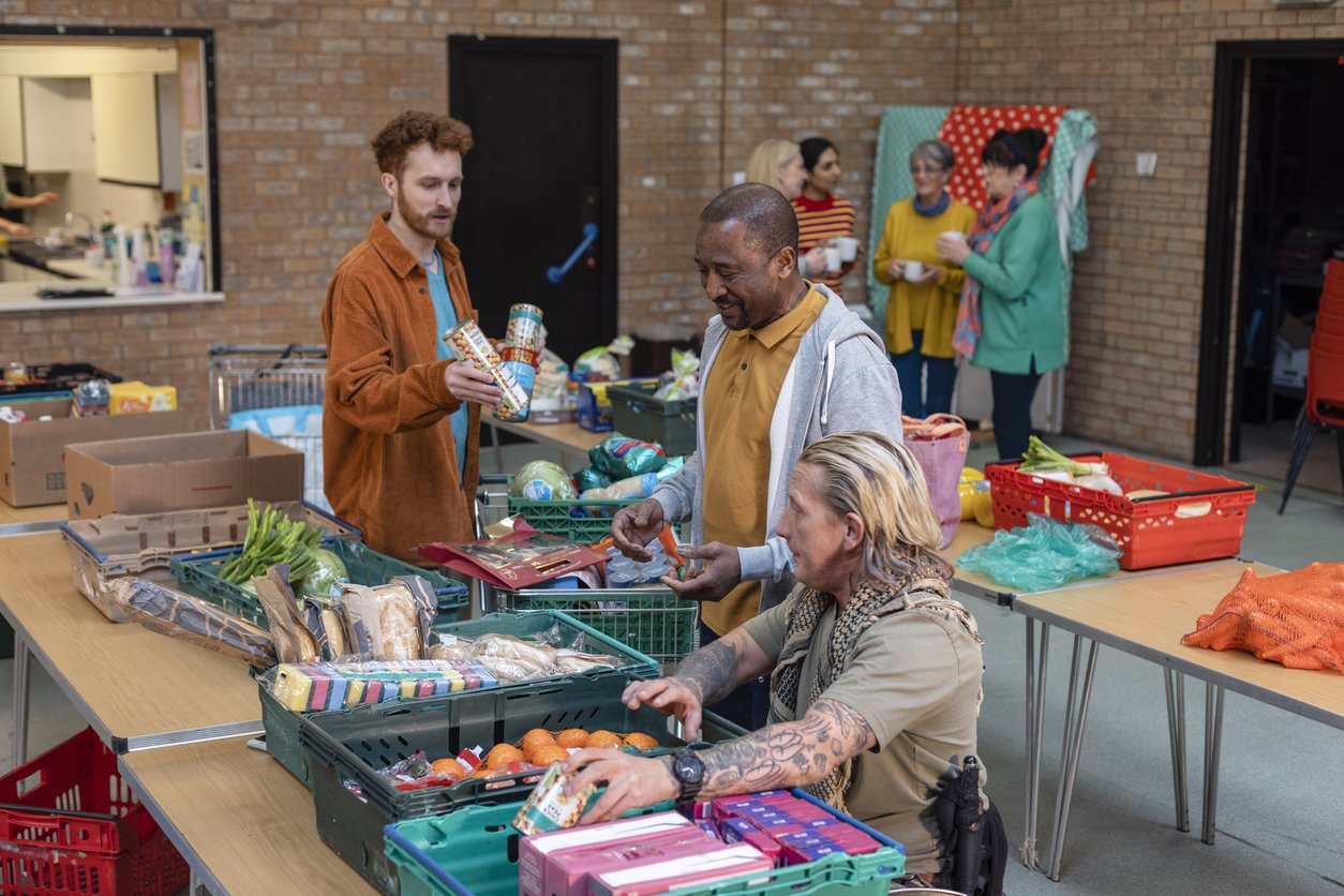 Male group of volunteers organising food donations onto tables at a food bank in the North East of England. They are working together, setting up sections of the room in a church. A group of women are drinking hot drinks out of focus in the background.
