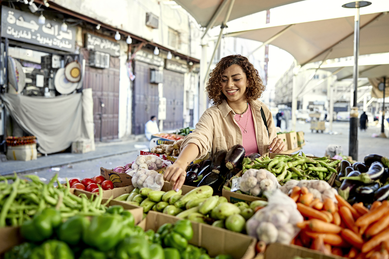 Waist-up view of smiling Middle Eastern woman in casual attire selecting zucchini from variety of vegetables in retail display under protective umbrellas.