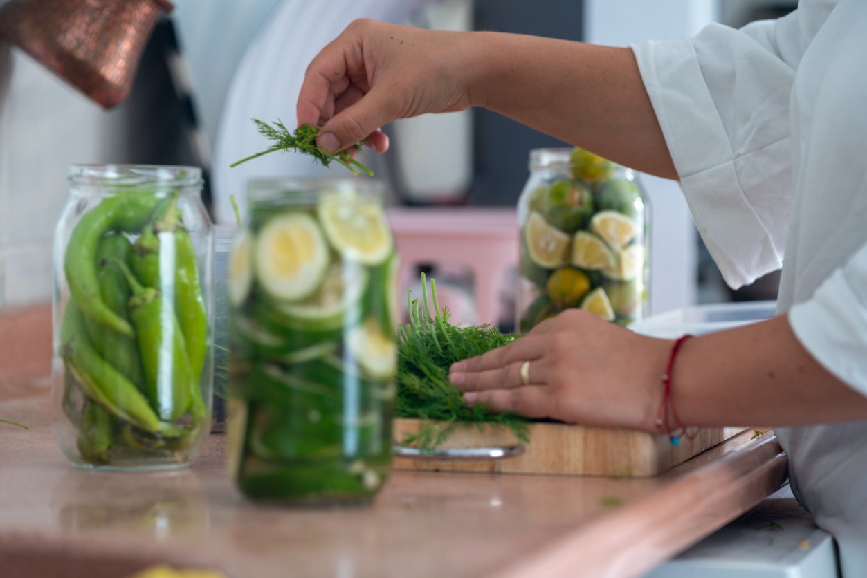 Photo of woman hand preparing fruit and vegetables for seasoning. Limes, lemons, oranges and green peppers are seen in jars. Shot from a low angle viewpoint with a full frame mirrorless camera in kitchen.
