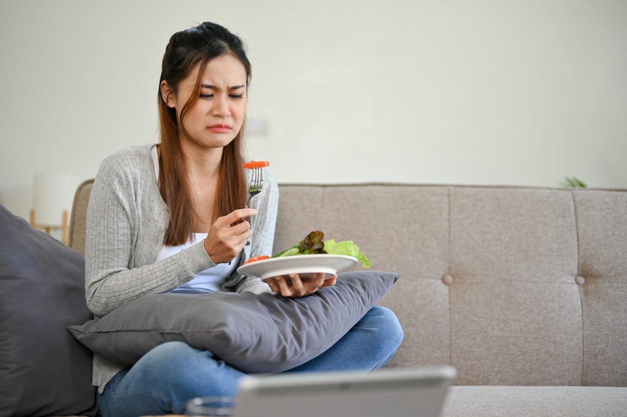 Unhappy and sad young Asian female is on diet, trying to eat fresh vegetables or salad to lose her weight, sitting on her couch in living room.