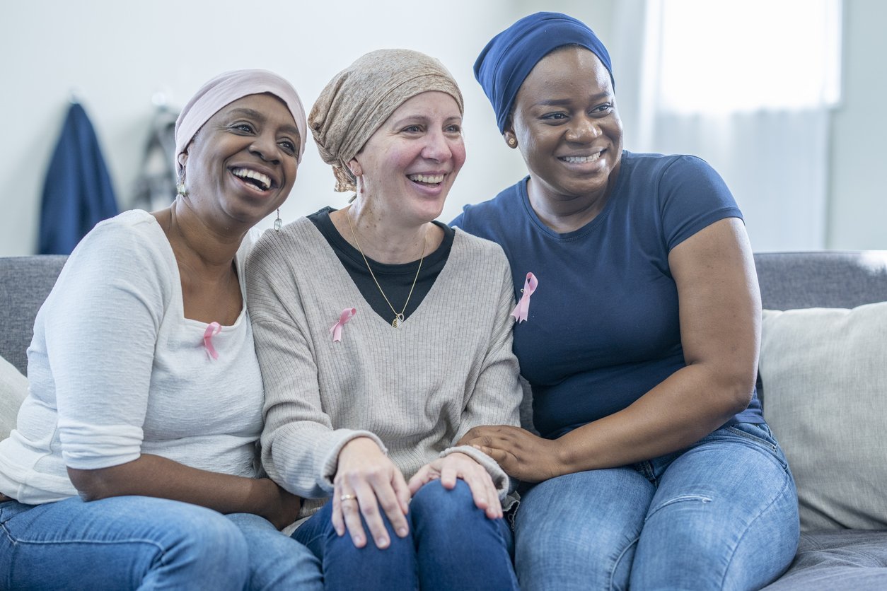 A group of three women fighting cancer, huddle in closely to one another on a sofa as they support each other.  They are each dressed casually, have head scarves on to keep them warm and are wearing pink Breast Cancer awareness ribbons.