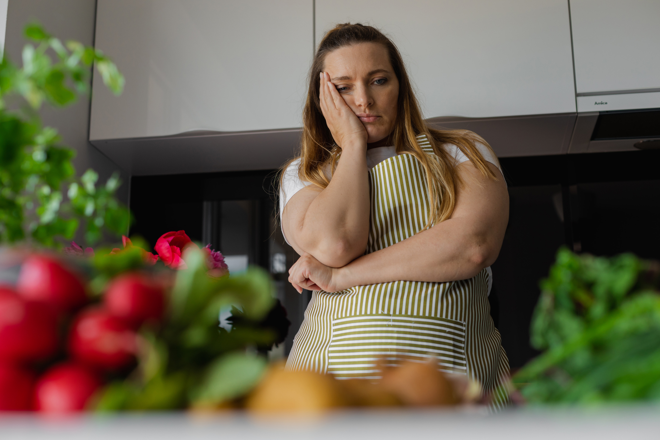 Tired, sad and bored plus size blond woman holding head in hand, thinking what to cook, have no idea. Diet and healthy eating with vegetables. Home cooking, looking for recipe. Greenery on foreground