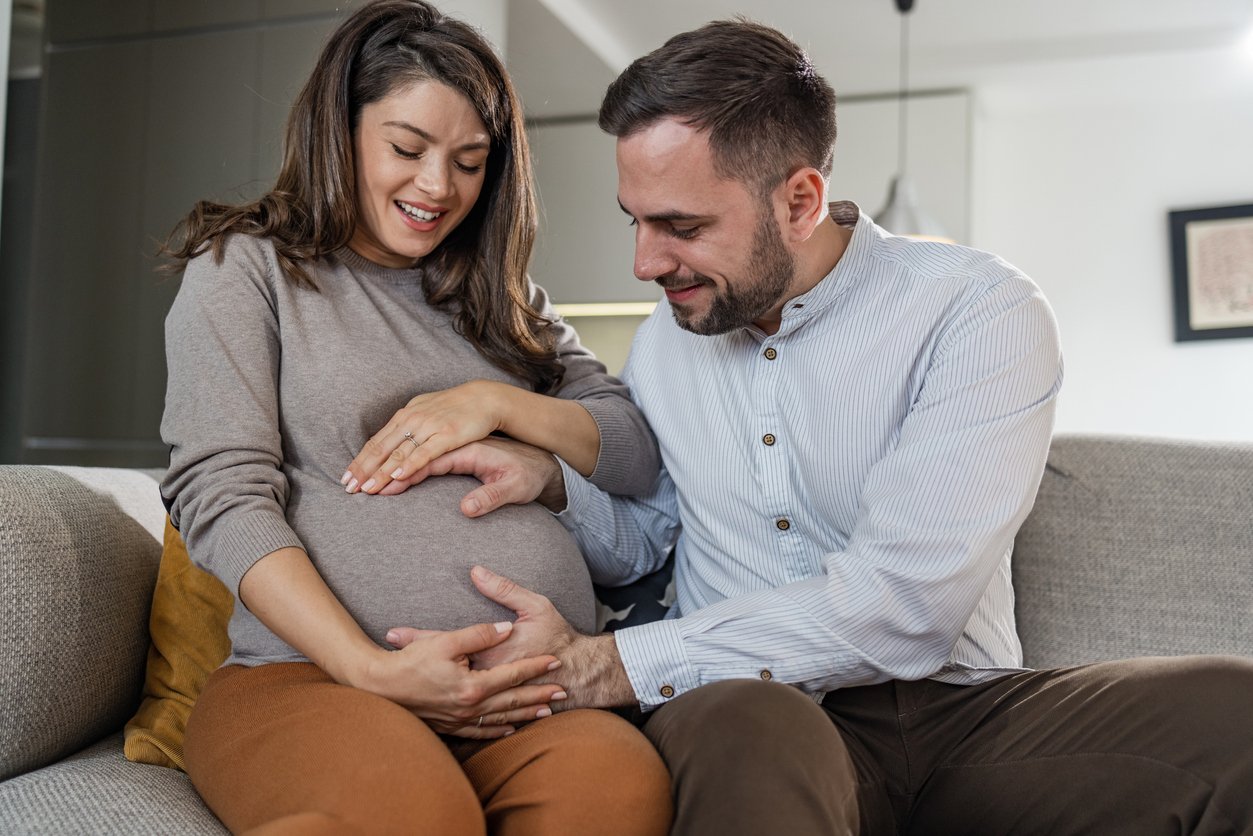 Mid-adult heterosexual pregnant couple sitting on the sofa in the living room. A man is gently holding a woman's belly.