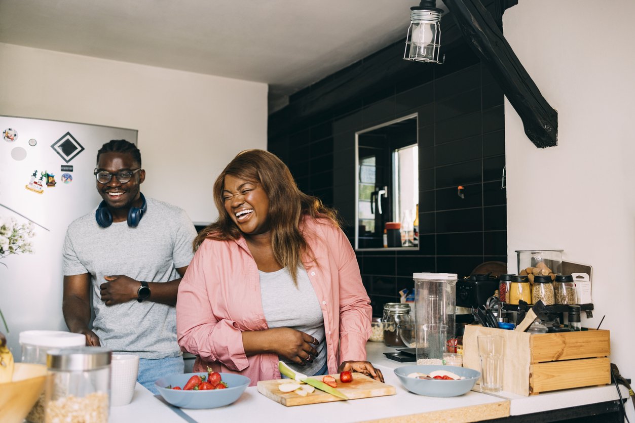 Улыбающаяся афроамериканская пара стоит на кухне и веселится, готовя здоровую еду.