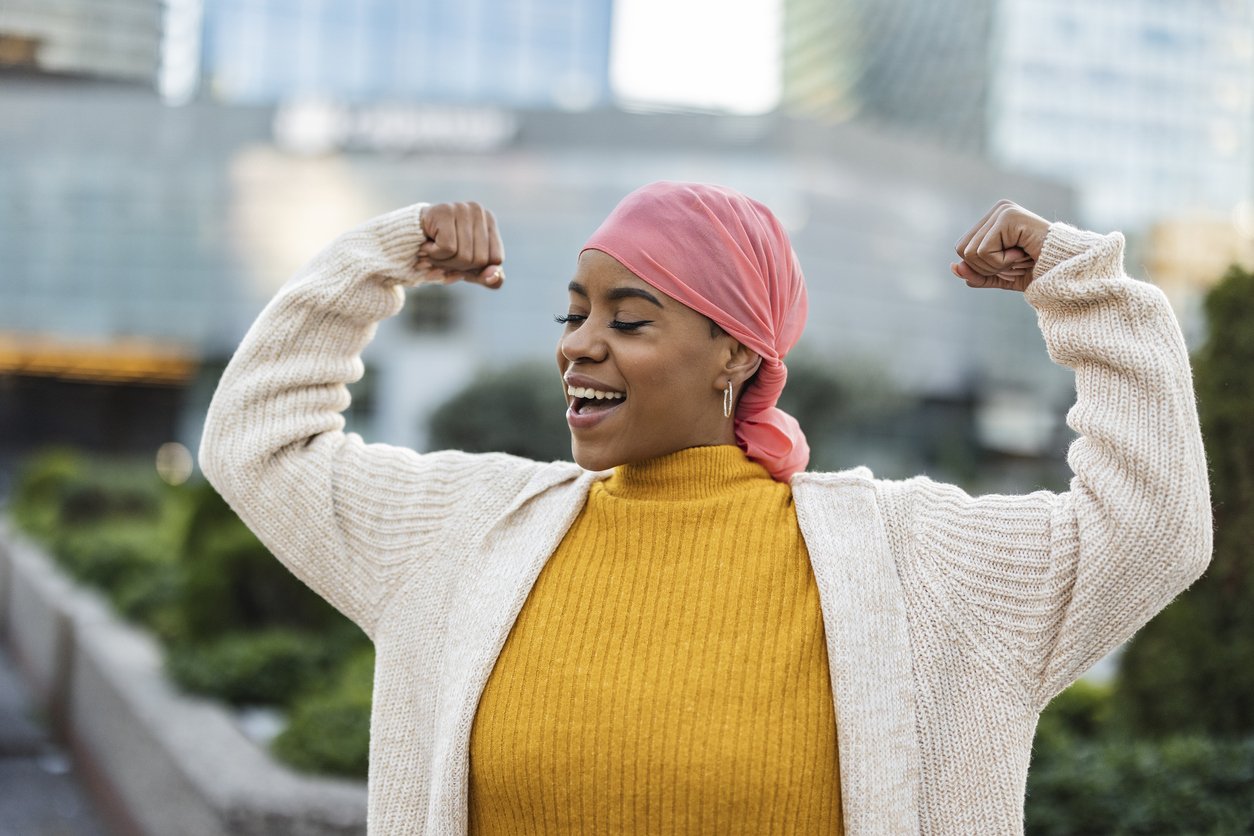 Latina woman, fighting breast cancer, wears a pink scarf, and clenches her arms as a survivor fighter