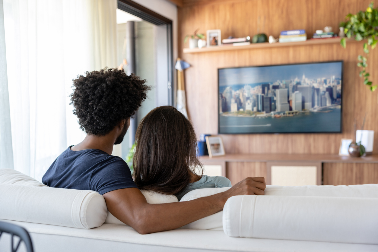 Black couple watches a documentary on a TV with their backs to the camera.