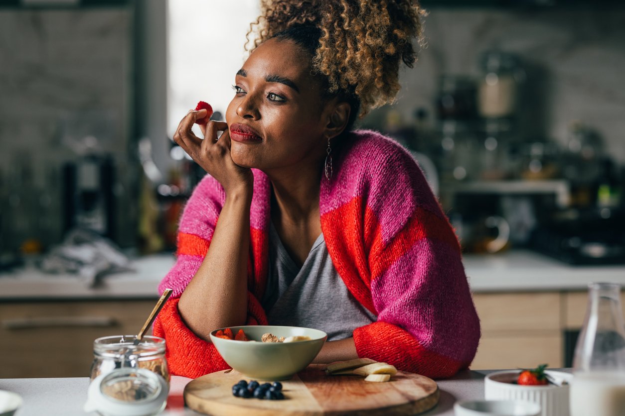 A pensive smiling African-American female contemplating while eating fresh organic strawberries. She is sitting in the kitchen.