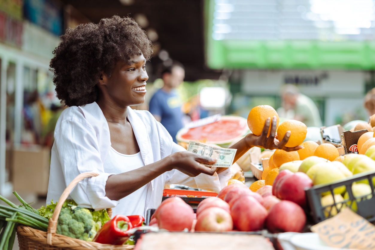 Smiling young woman holding a basket full of groceries on the farmer's market and choosing fresh vegetables. She is paying with cash