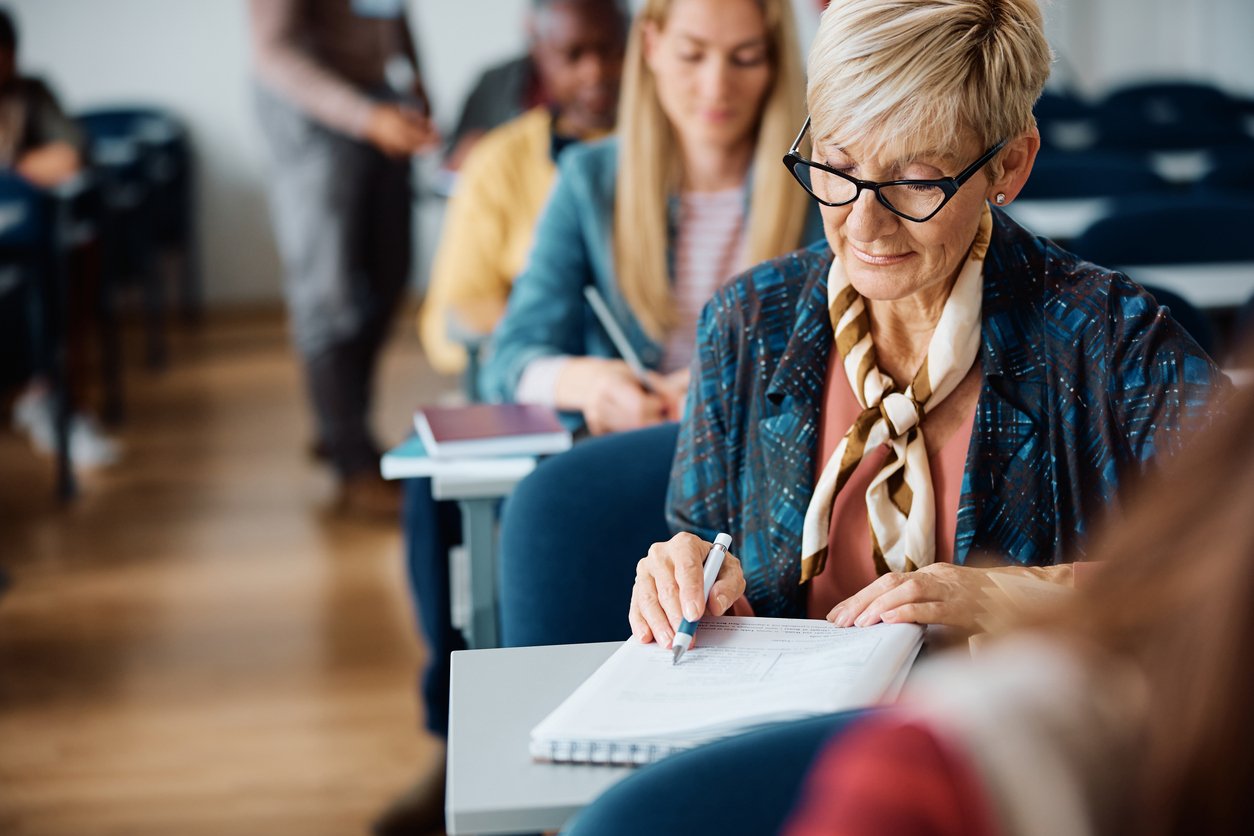 Mature female student taking notes while attending a course in lecture hall.