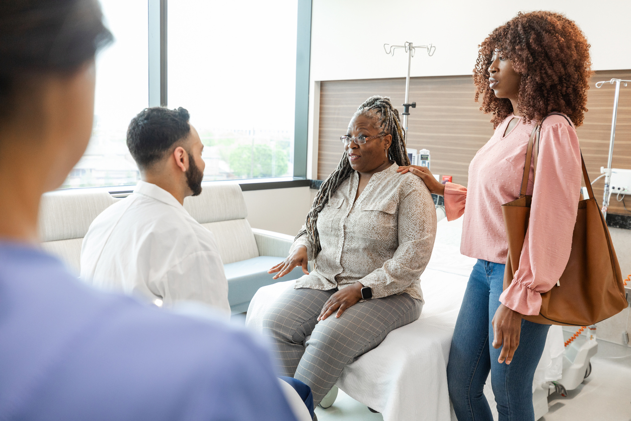 As an unrecognizable nurse watches, the serious mature adult woman and her mid adult daughter talk to the unrecognizable male emergency room doctor.