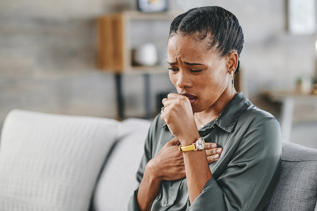 Woman sitting on sofa at home, frowning and pressing on chest, coughing into palm