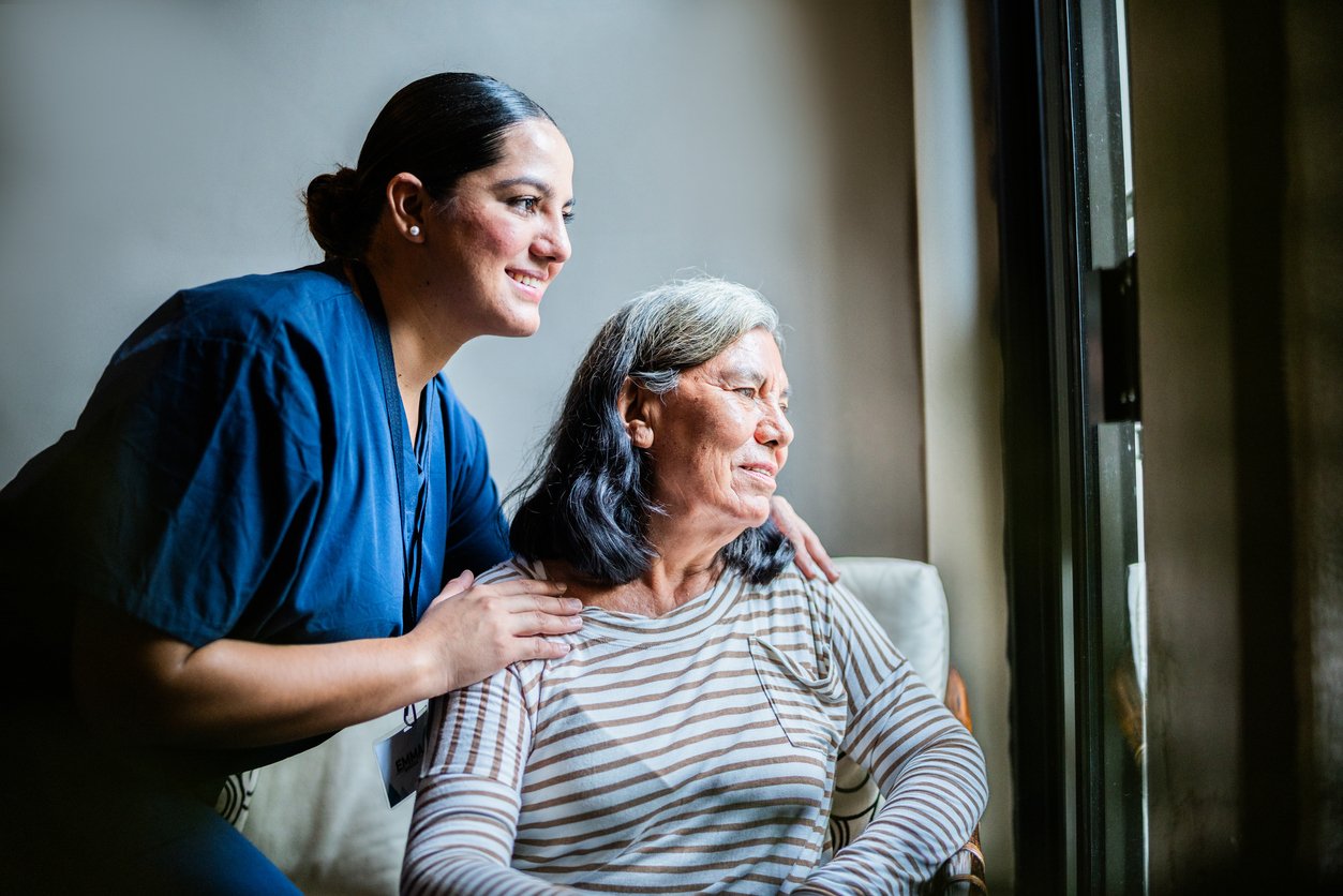 Home caregiver and senior woman looking through window at home