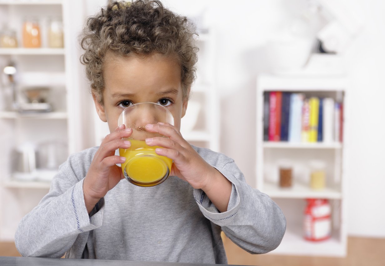 A close-up of a biracial toddler drinking orange juice in the kitchen.