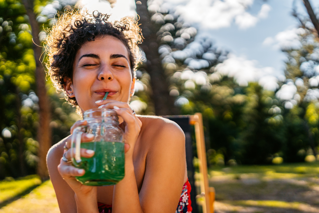 Beautiful young woman in a red dress dancing drinking green juice from a jar in the bar outdoors.