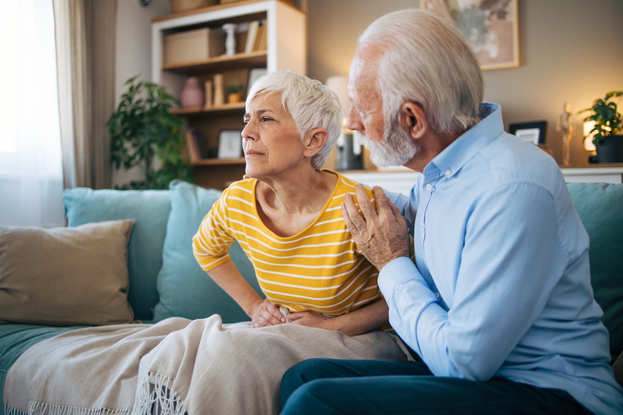 Husband taking care about his sick wife. Senior heterosexual couple sitting on the sofa. Woman has painful facial expresion because of stomachache.