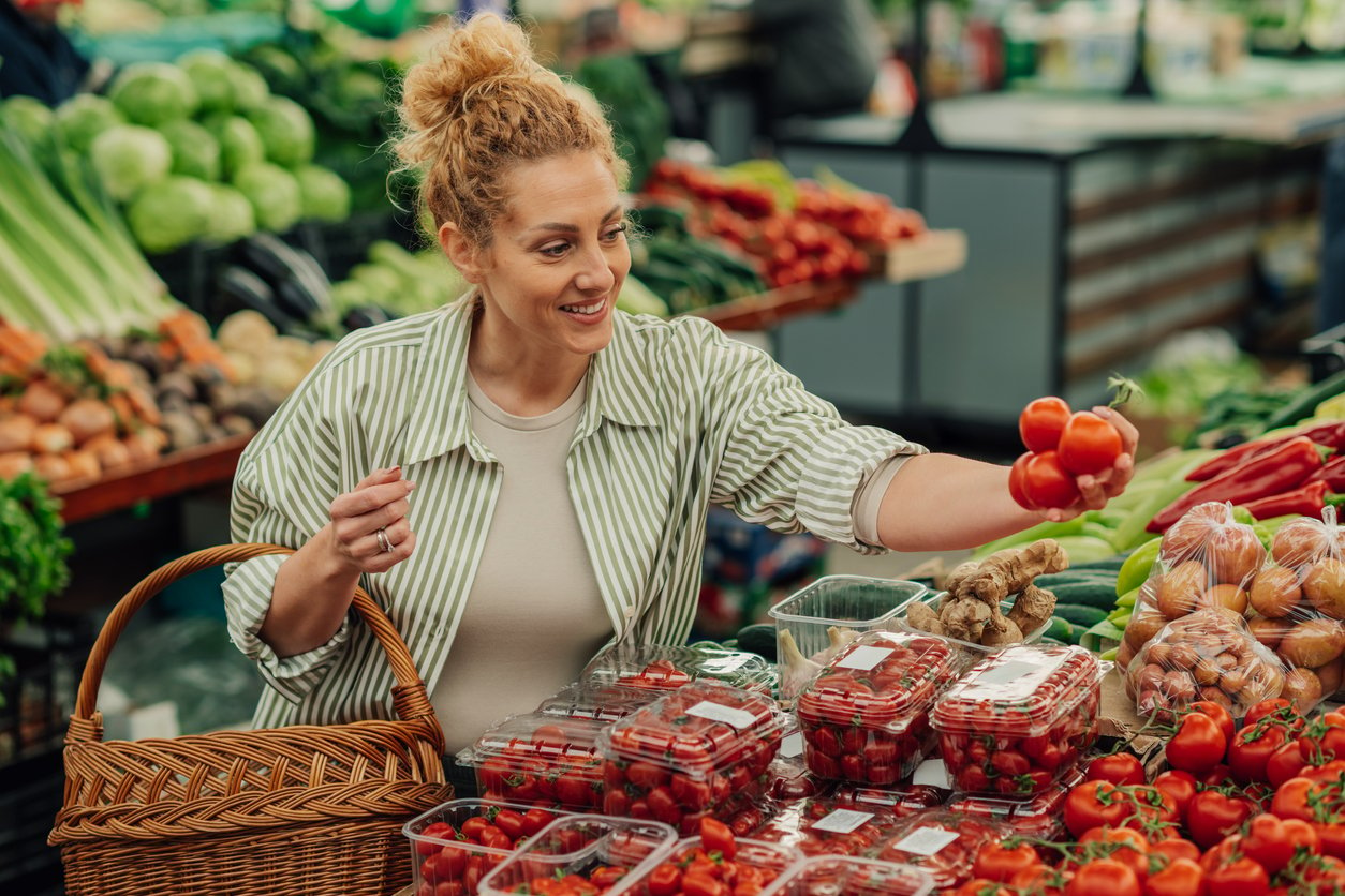 Smiling female customer standing near market stall at farmers market with wicker shopping basket in hands and choosing organic tomatoes. Portrait of a shopper woman buying healthy food at green market