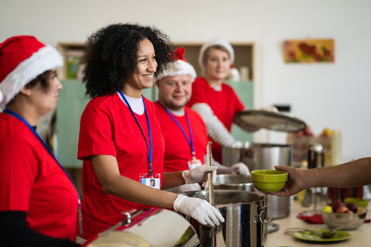 A woman is smiling at someone holding out a bowl to receive some food, standing in a line with more volunteers wearing christmas hats and red shirts while they work in a soup kitchen during the festive season, bringing hot and healthy food for deprived people