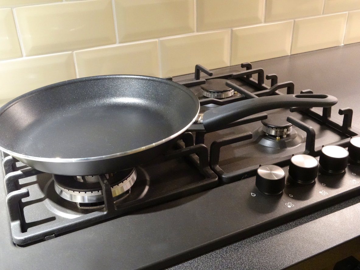 Photo showing a black, aluminium non-stick frying pan, which is sitting on top of an integrated gas cooker hob, on a granite worktop / countertop.  The gas rings are switched off, after being cleaned.