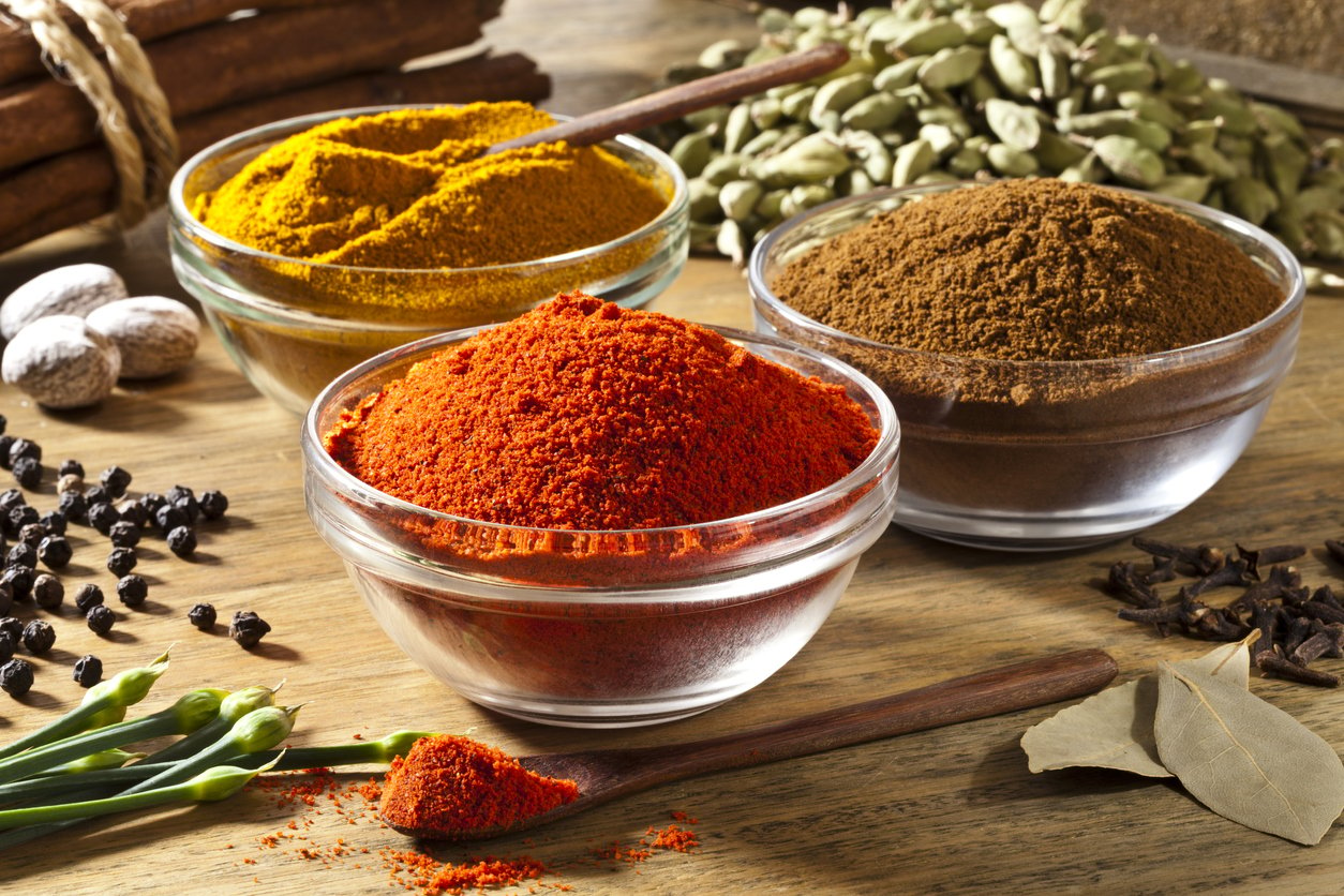 Three transparent glass bowls filled with ground spices are arranged in a triangle shape on a rustic wood table. Other spices like black pepper, nutmeg, cinnamon sticks, cardamom, clove and bay leaf are around the bowls. In the foreground there is a little wood spoon filled with paprika. Main light comes from the top-left giving a nice shadow to the picture. Very saturated and colorful image taken with DSRL Canon EOS 5D Mk II.