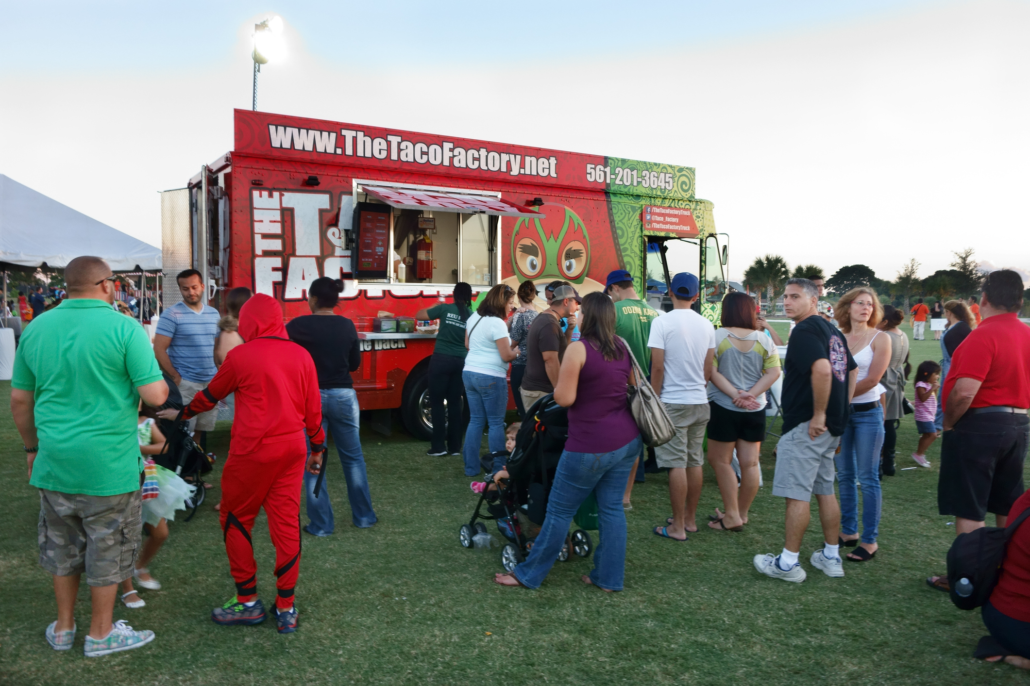 People socialize and order food in front of a Tacos Factory food truck at a public event