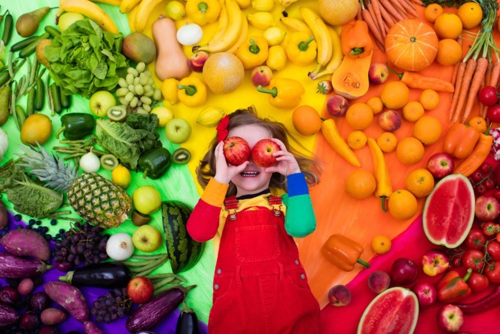 Young girl using apples for eyes surrounded by a rainbow of fruits and vegetables