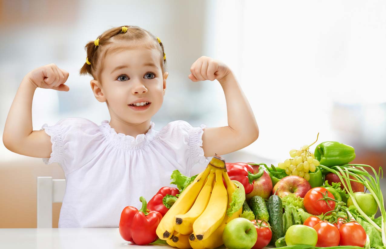 young girl holding up strong arms behind fruits and veggies