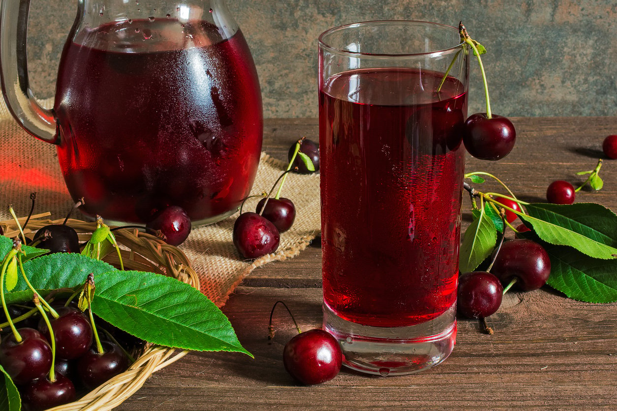 cold cherry juice in a glass and pitcher with cherries inside on wooden table with ripe berries in wicker basket