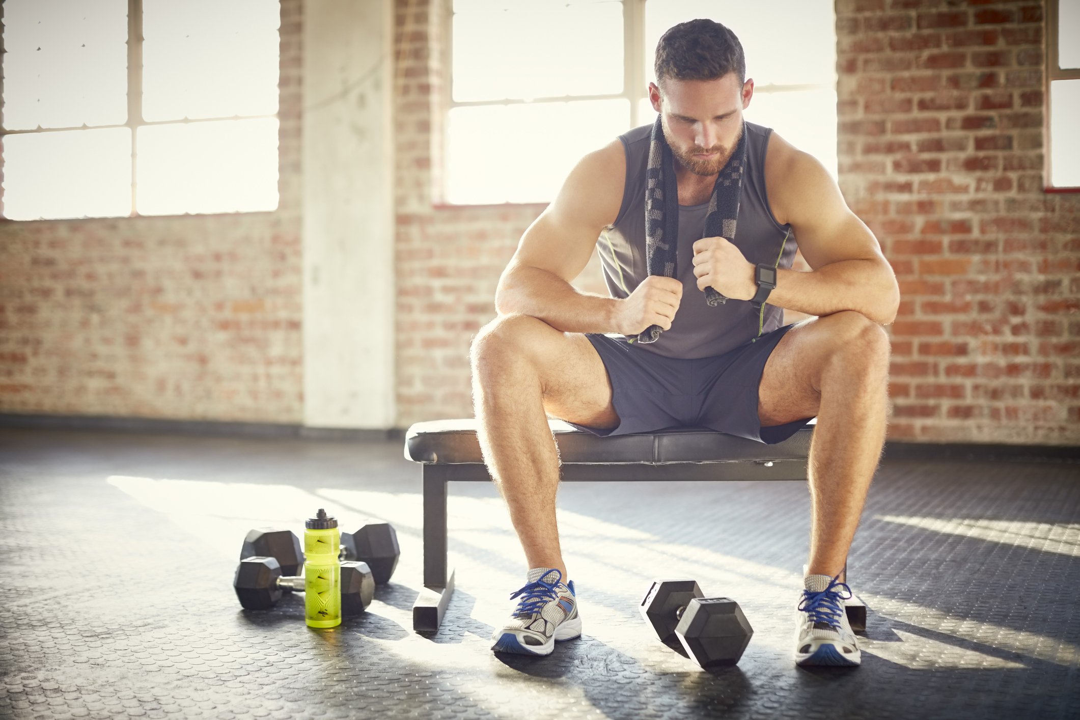 Full length of tired young man sitting on bench in gym. Attractive male is with towel around neck after workout. He is resting in health club.