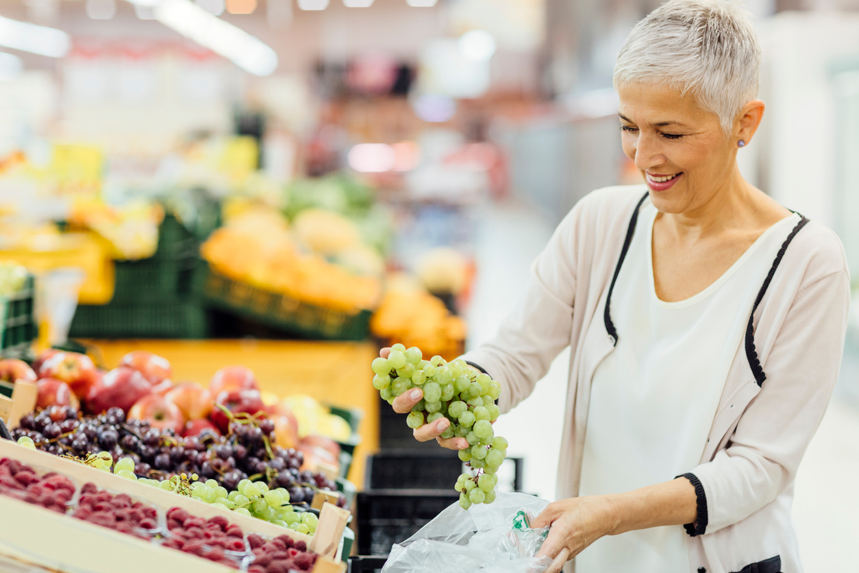 面帶微笑的成熟女性在當地超市購物。她正在選購日用品，挑選水果、葡萄。