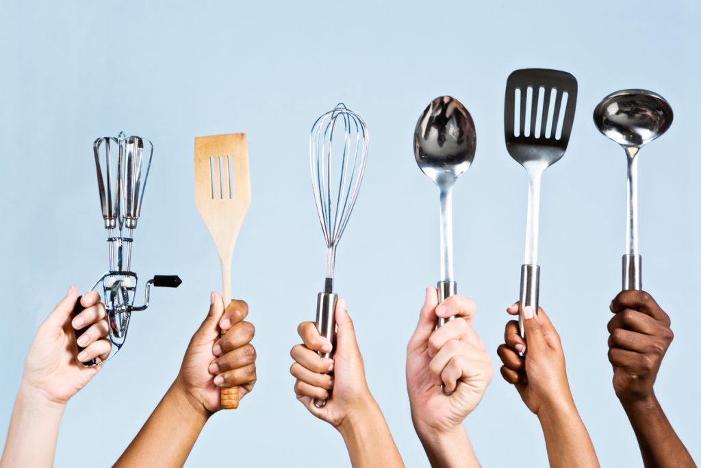 Six mixed hands hold up a kitchen ladle, a spoon, a beater, a whisk, an egg lifter and a wooden spatula. They're ready to cook almost anything!