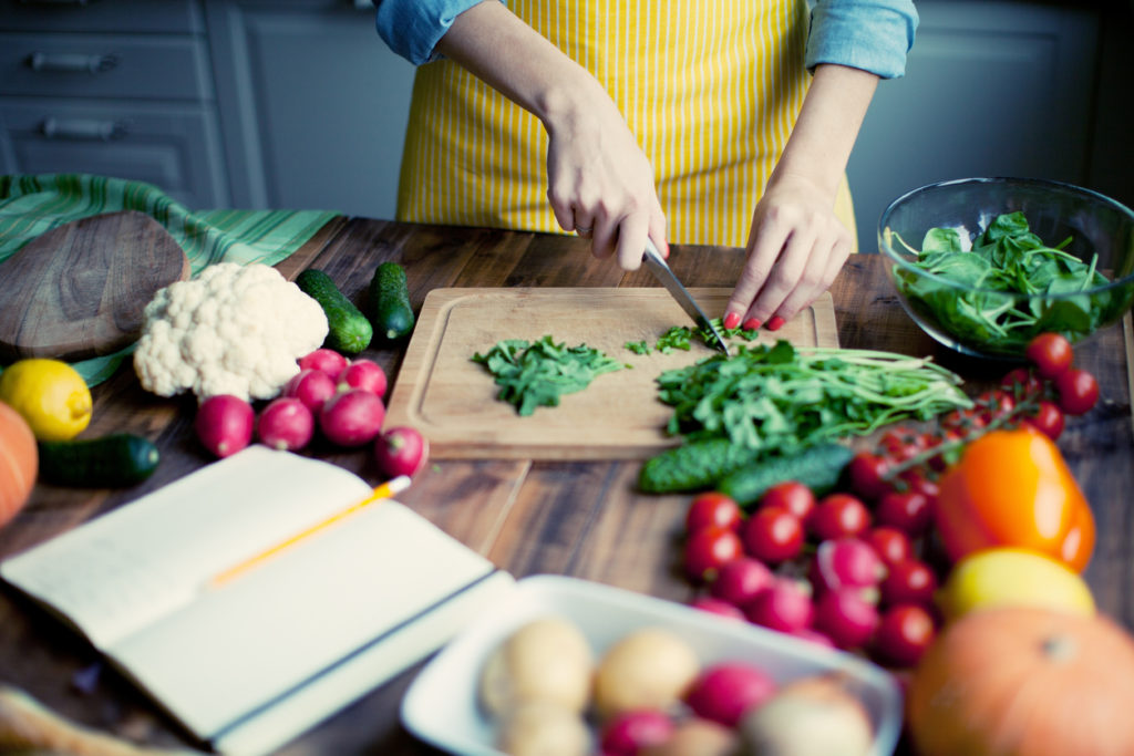 Woman chopping green vegetables on a cutting board in the kitchen, surrounded by other fruits and vegetables