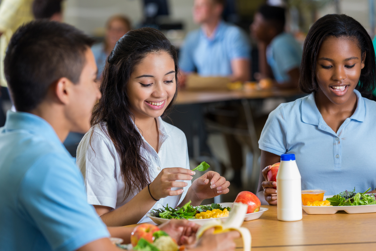 Diverse male and female high school friends eat lunch together in school cafeteria.