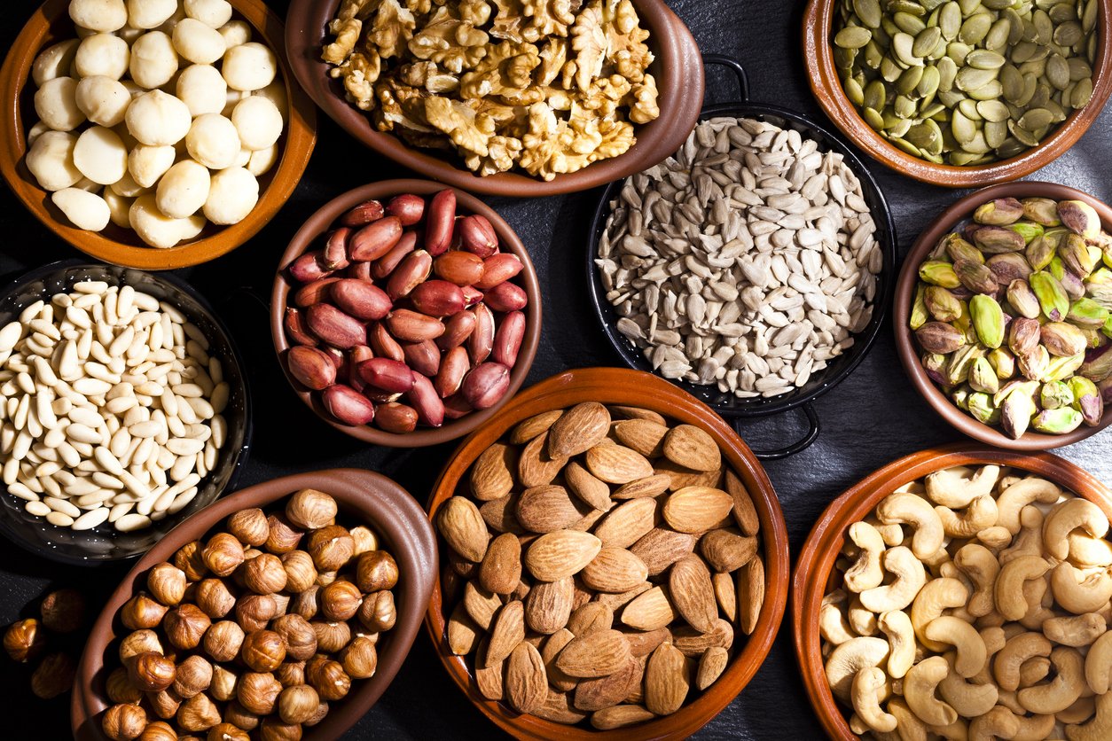 Top view of a black table filled with a large assortment of nuts like pistachios, hazelnut, pine nut, almonds, pumpkin seeds, peanuts, cashew and walnuts. Nuts are in brown bowls. Predominant color is brown. DSRL studio photo taken with Canon EOS 5D Mk II and Canon EF 100mm f/2.8L Macro IS USM