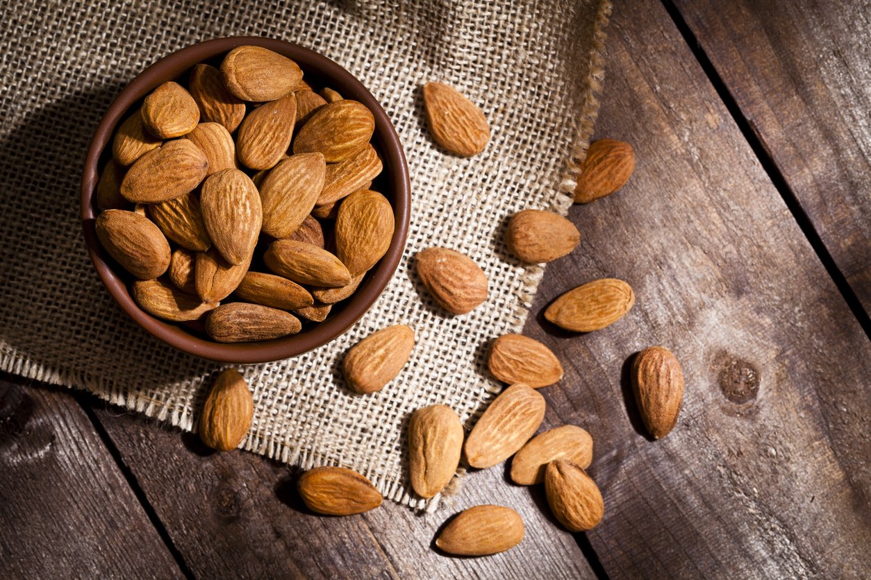 Top view of a brown bowl filled with organic almonds shot on rustic wood table. Some almonds are out of the bowl on a burlap. Predominant color is brown. DSRL studio photo taken with Canon EOS 5D Mk II and Canon EF 100mm f/2.8L Macro IS USM