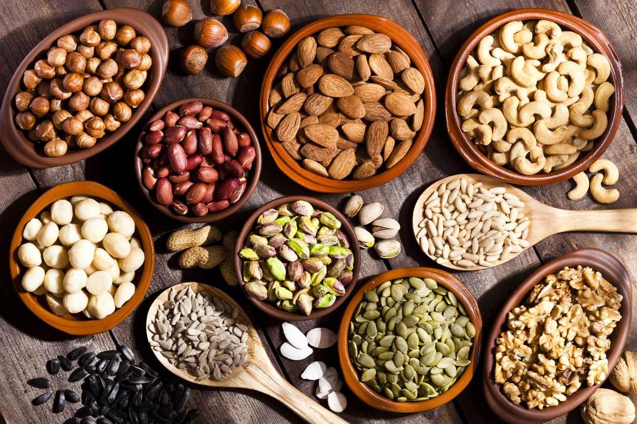 Top view of a rustic wood table filled with a large assortment of nuts like pistachios, hazelnut, pine nut, almonds, pumpkin seeds, peanuts, cashew and walnuts. Some nuts are in brown bowls and wooden spoon and others directly on the table. Predominant color is brown. DSRL studio photo taken with Canon EOS 5D Mk II and Canon EF 100mm f/2.8L Macro IS USM