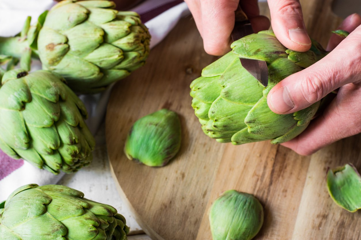 Man's hand holding knife and peeling fresh artichokes, preparing for cooking, cutting board, top view