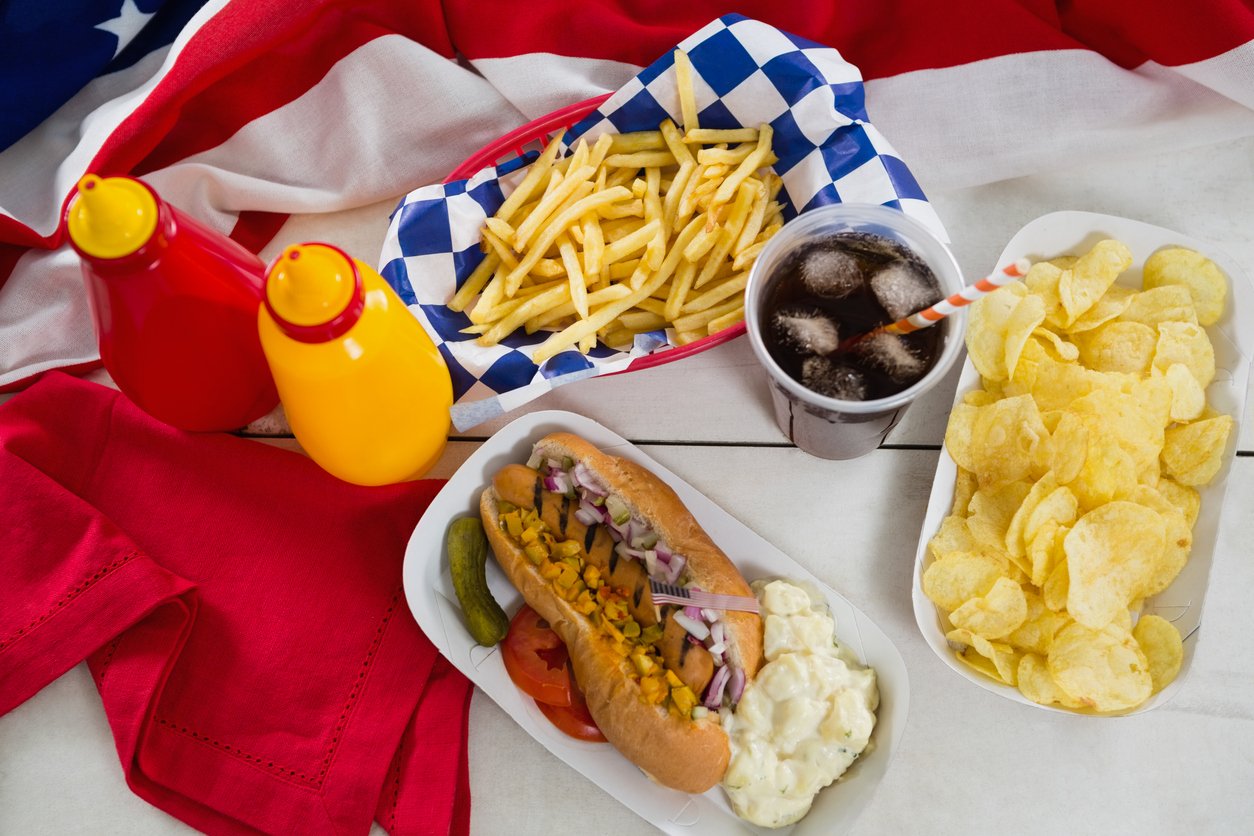 Hot dog with other unhealthy foods like french fries, soda, and chips next to American flag