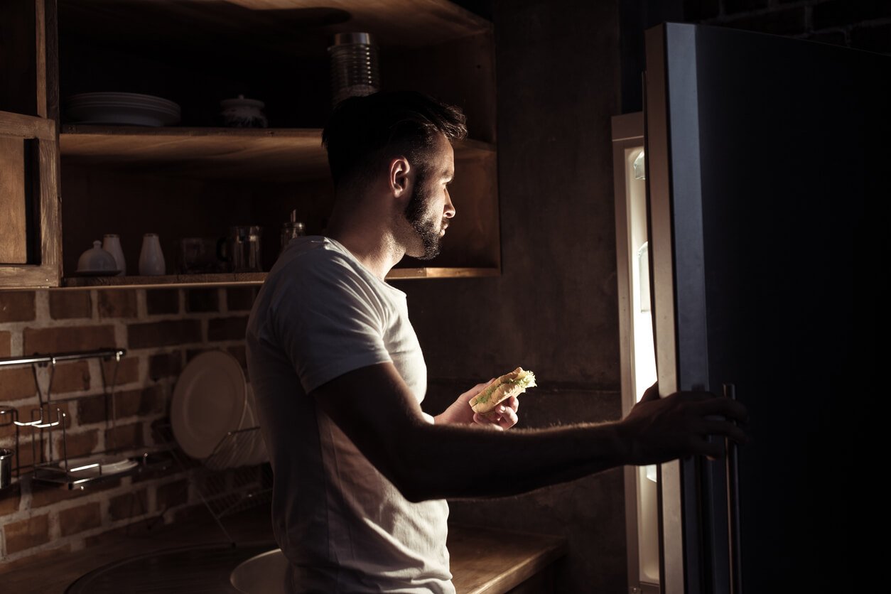 young man in pyjamas eating and looking at refrigerator at night