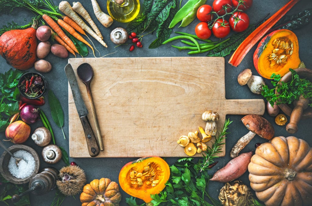 An empty cutting board with a myriad of fresh fall vegetables surrounding it