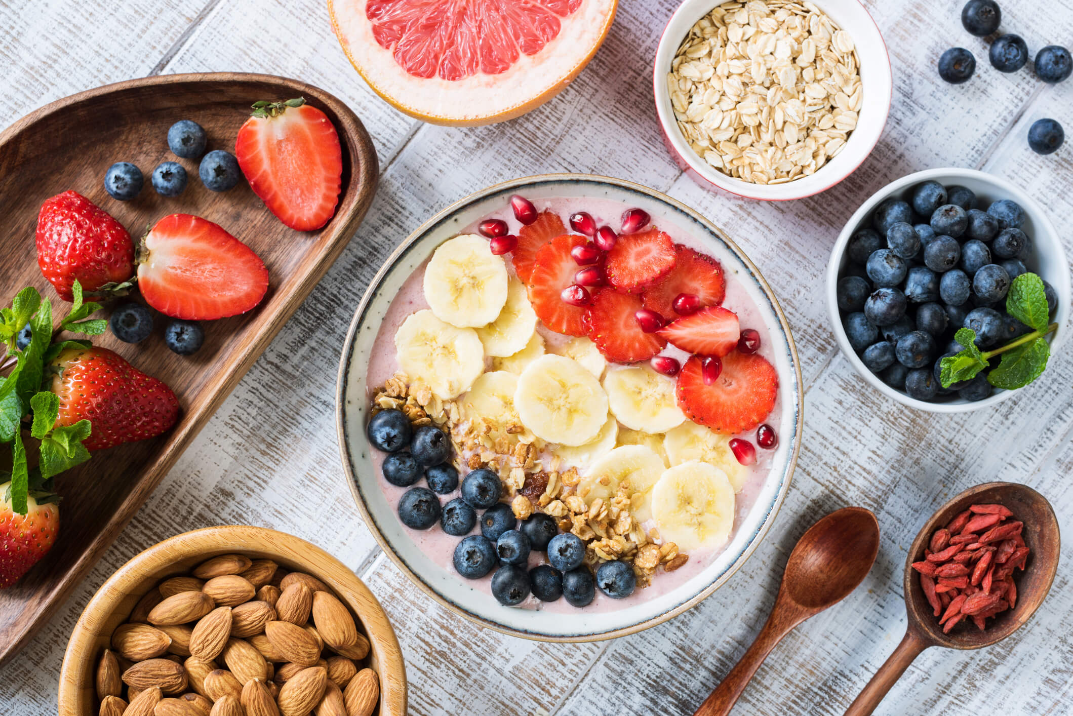  A wooden table with a bowl of smoothie topped with fruit and granola, and other bowls of fruit, nuts, and seeds.